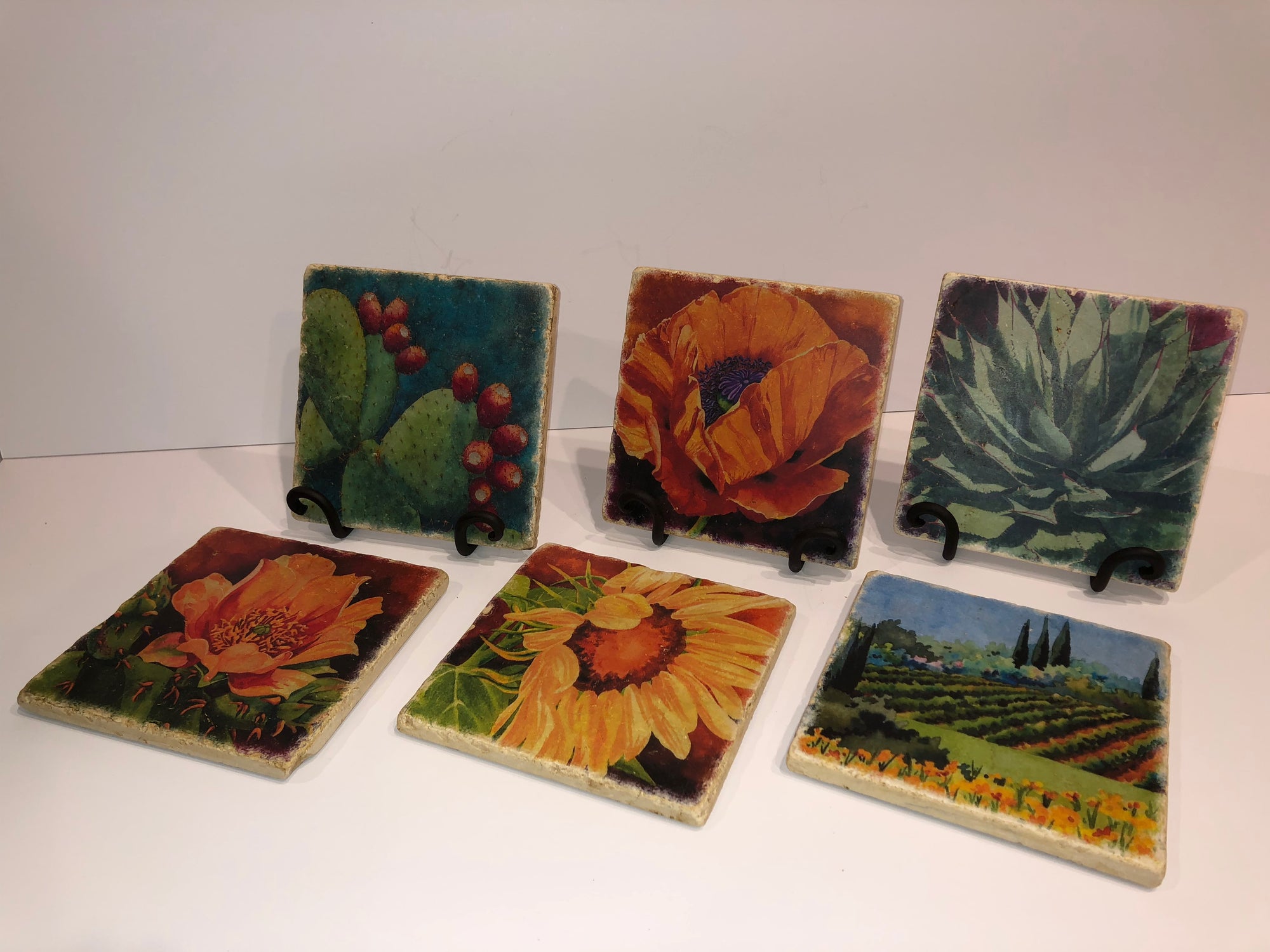 An assortment of Heidi Rosner trivets. The Fruit of the Opuntia, Brilliant Bloom, and Desert Lotus trivets are shown on iron stands. Prickly Pear 2, Sunflower 1, and Vineyard I trivets are shown flat on the table.